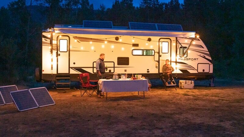 A recreational vehicle (RV) parked in a forest at night with its interior and exterior lights turned on. The RV is powered by Point Zero Energy's portable solar panels and portable power station, which can be seen placed on the ground beside the vehicle. 