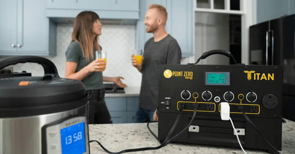 On a kitchen counter, the Point Zero Energy Titan Solar Power Station is shown with an Instapot connected to it. In the background, a husband and wife are seen enjoying a glass of orange juice.