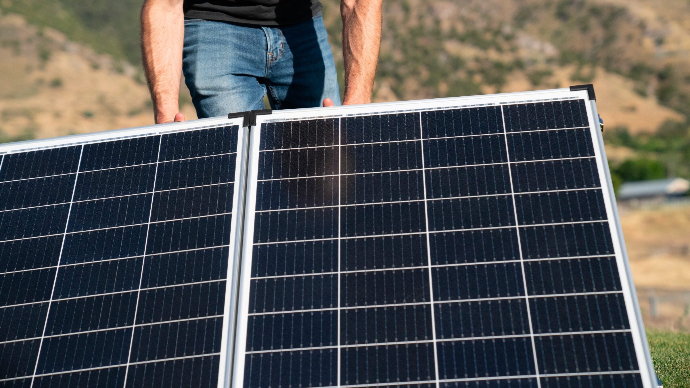 A man wearing a black shirt and blue jeans is setting up solar panels on a metal frame outside in the bright sunlight. 