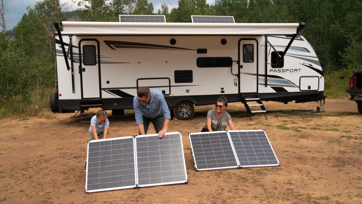 a family of three - a mom, dad, and their daughter - installing Point Zero Energy solar panels in front of their RV, which is parked in a wilderness area. The daughter is holding onto one of the solar panels while the parents secure it in place. The family is surrounded by trees, bushes, and mountains, suggesting they are enjoying a remote camping experience while also utilizing clean, renewable energy.